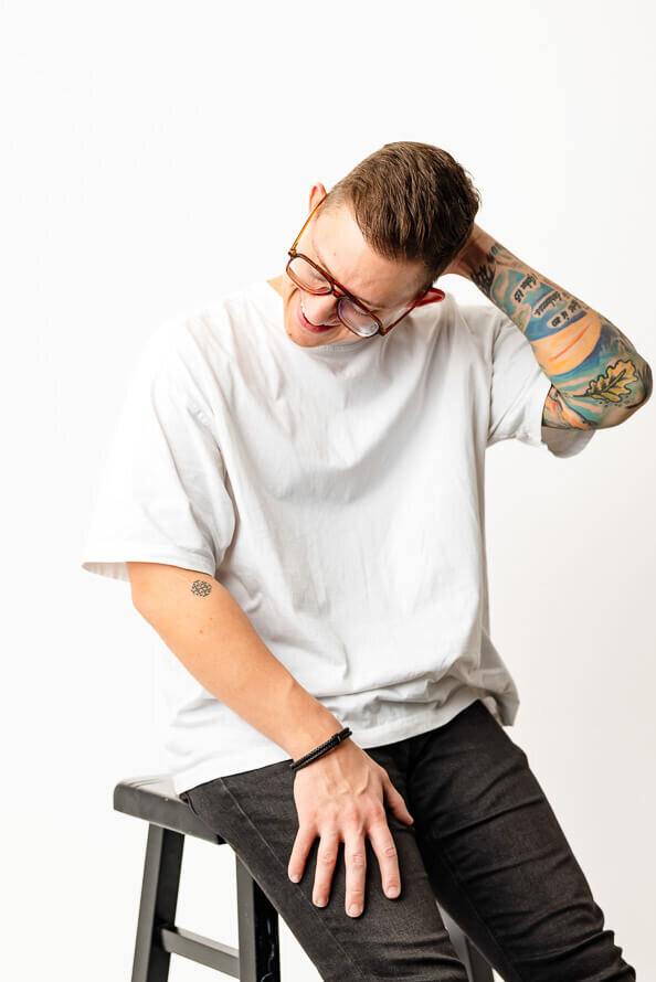 Man with colorful tattoos on arm puts hand behind head and looks down.