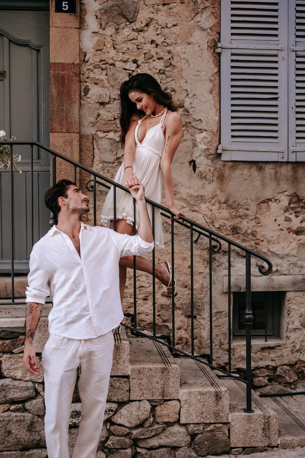 Woman at the top of the stairs looking down and holding onto her fiance as he stands in front of the staircase.