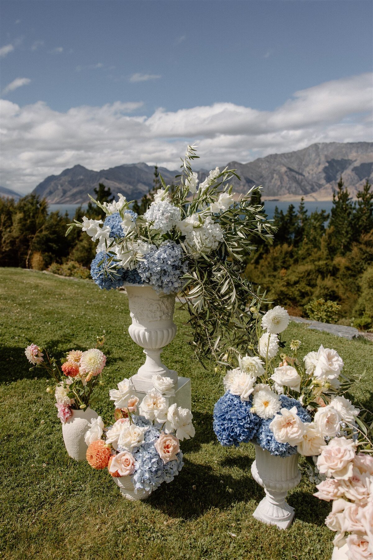 Romantic floral arrangements in soft blue, peach, and white tones, displayed in ceramic and stone vases on green grass with lake and mountain views behind.
