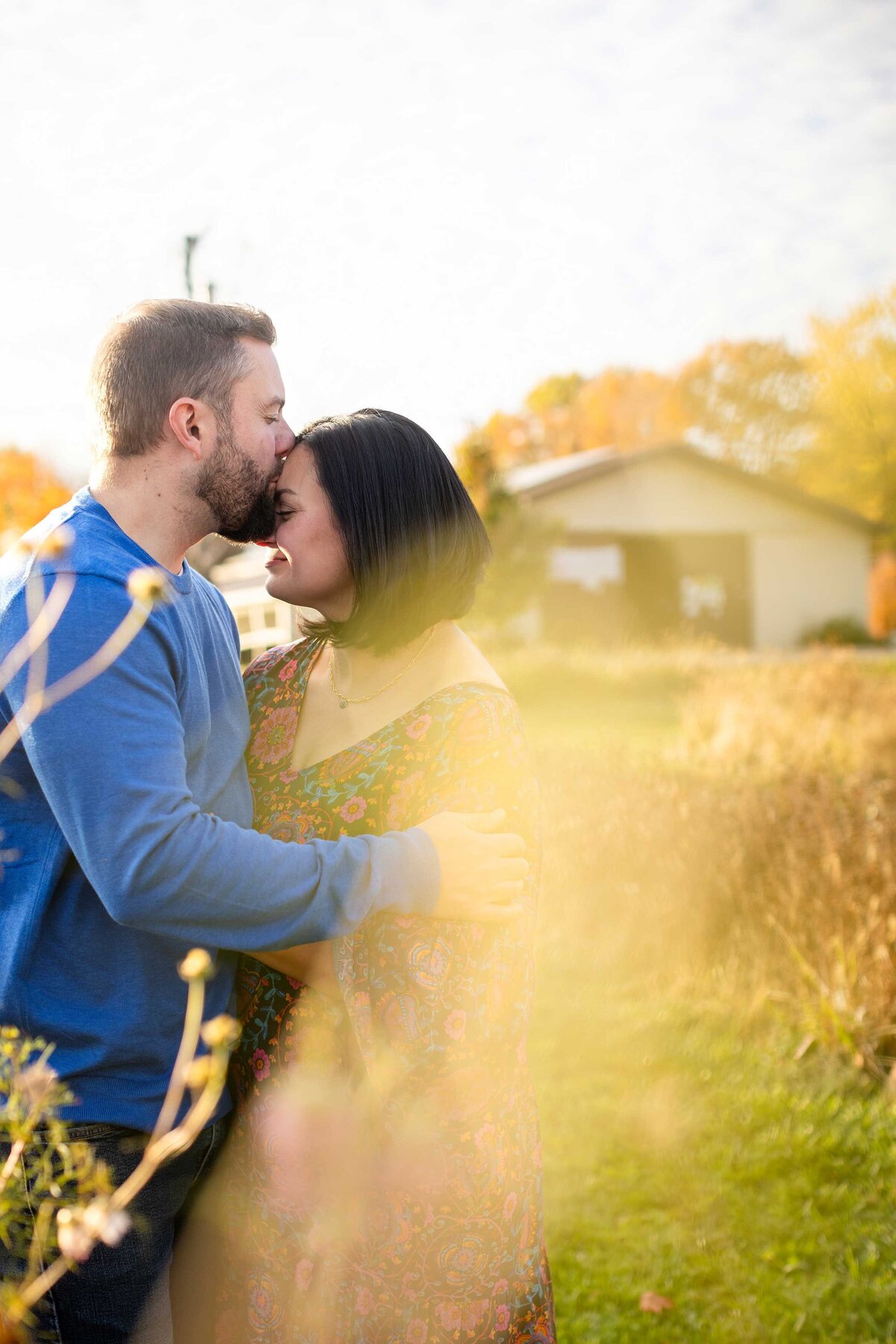 A romantic moment captured as a man in a blue shirt embraces a woman in a floral dress, kissing in a golden-lit field during an autumn sunset in Metro Detroit.