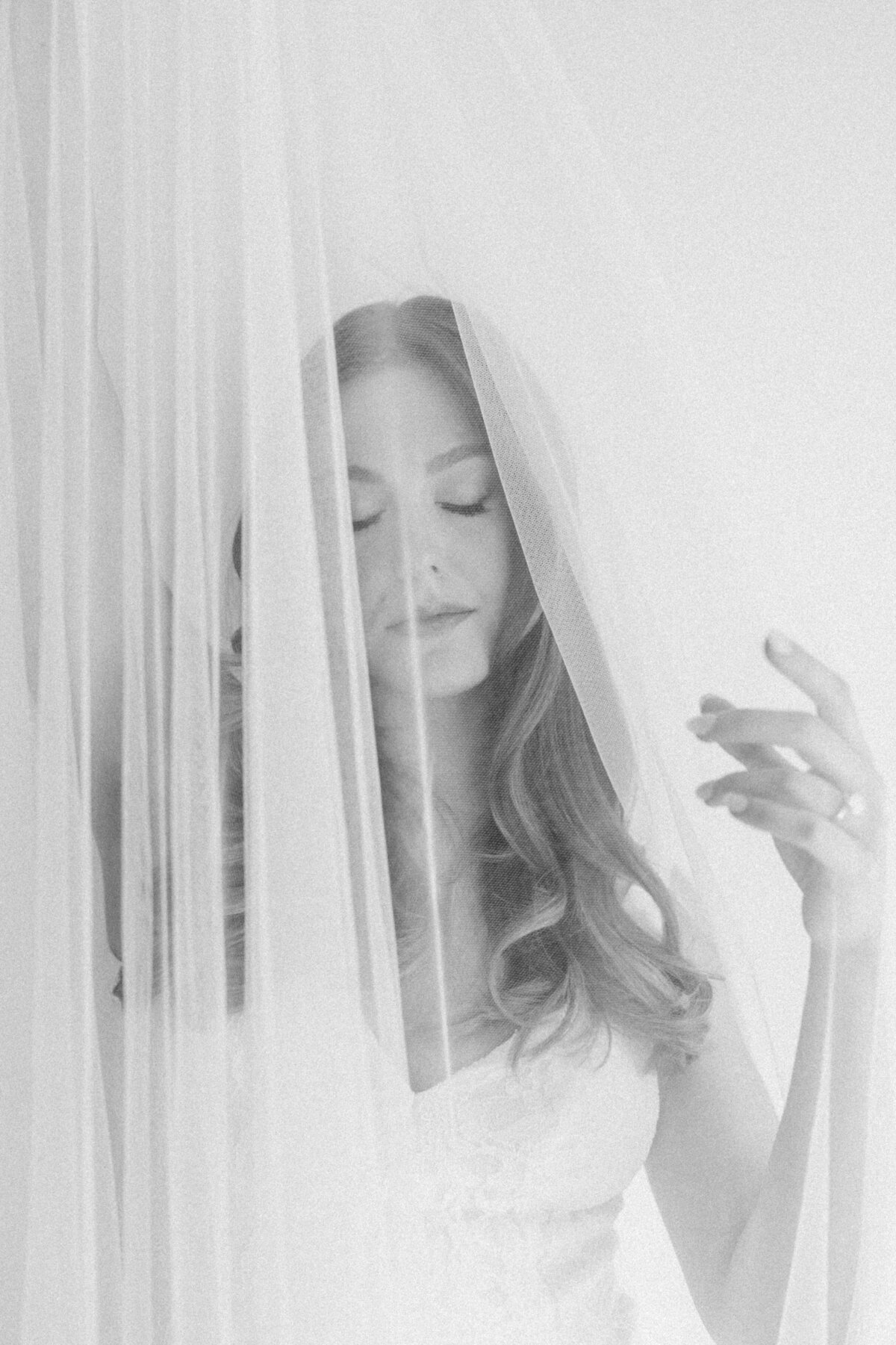 A delicate black-and-white image of a pregnant bride with eyes closed, enveloped in a flowing veil, creating a soft and ethereal effect