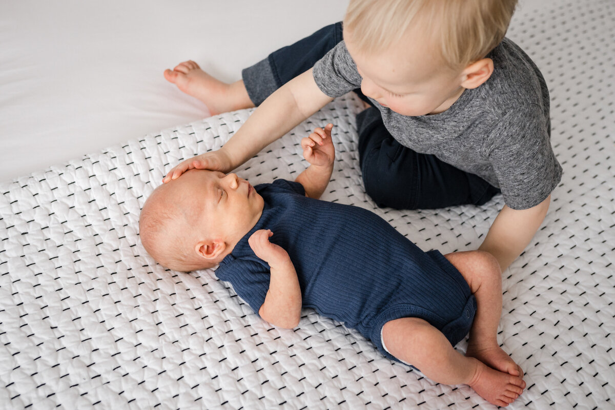 A toddler gently pats the head of a newborn baby lying on a textured white blanket.