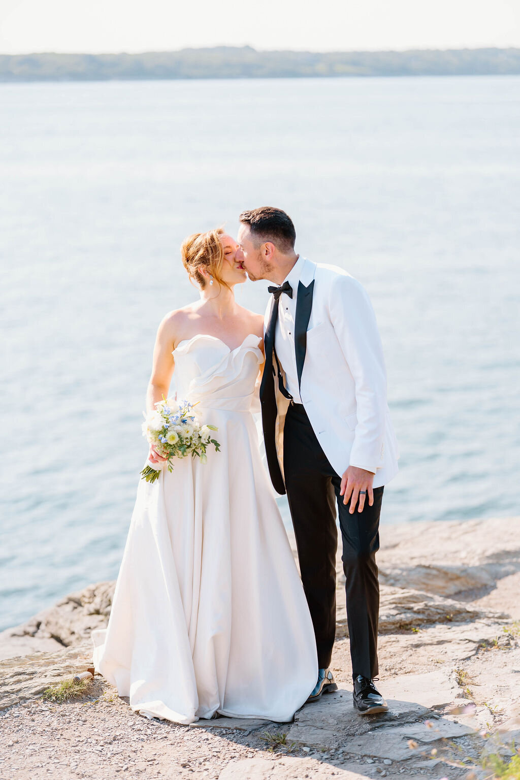 Rhode Island Elopement | A bride in a flowing white gown and a groom in a white tuxedo kiss on a rocky cliff by the sea. 