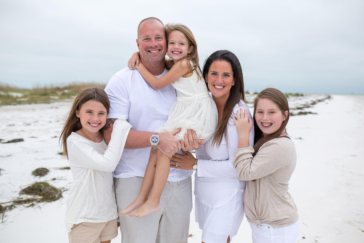 Father holding youngest daughter while wife and two other children hug them in a family portrait on the beach in Anna Maria Island, FL