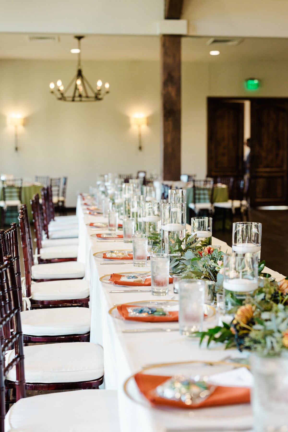 A detail shot of a head table at a wedding reception at the Laurel in Grapevine, Texas. The long, rectangular table has a long white tablecloth, place setting that have cutlery, orange napkins, and custom cookies on each plate as well as many candles and lots of florals and greenery as centerpieces. Other similar circular tables can be seen in the background.