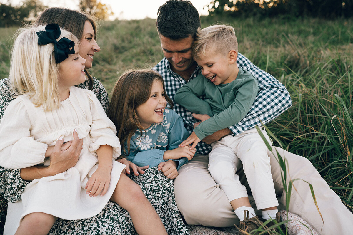 Family of four sitting in tall grass, laughing and enjoying a carefree moment during their photo session