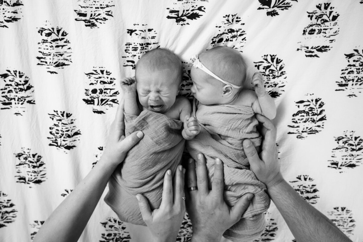 A black and white image of newborn twins lying on a patterned bedspread, each held securely by their parents' hands.