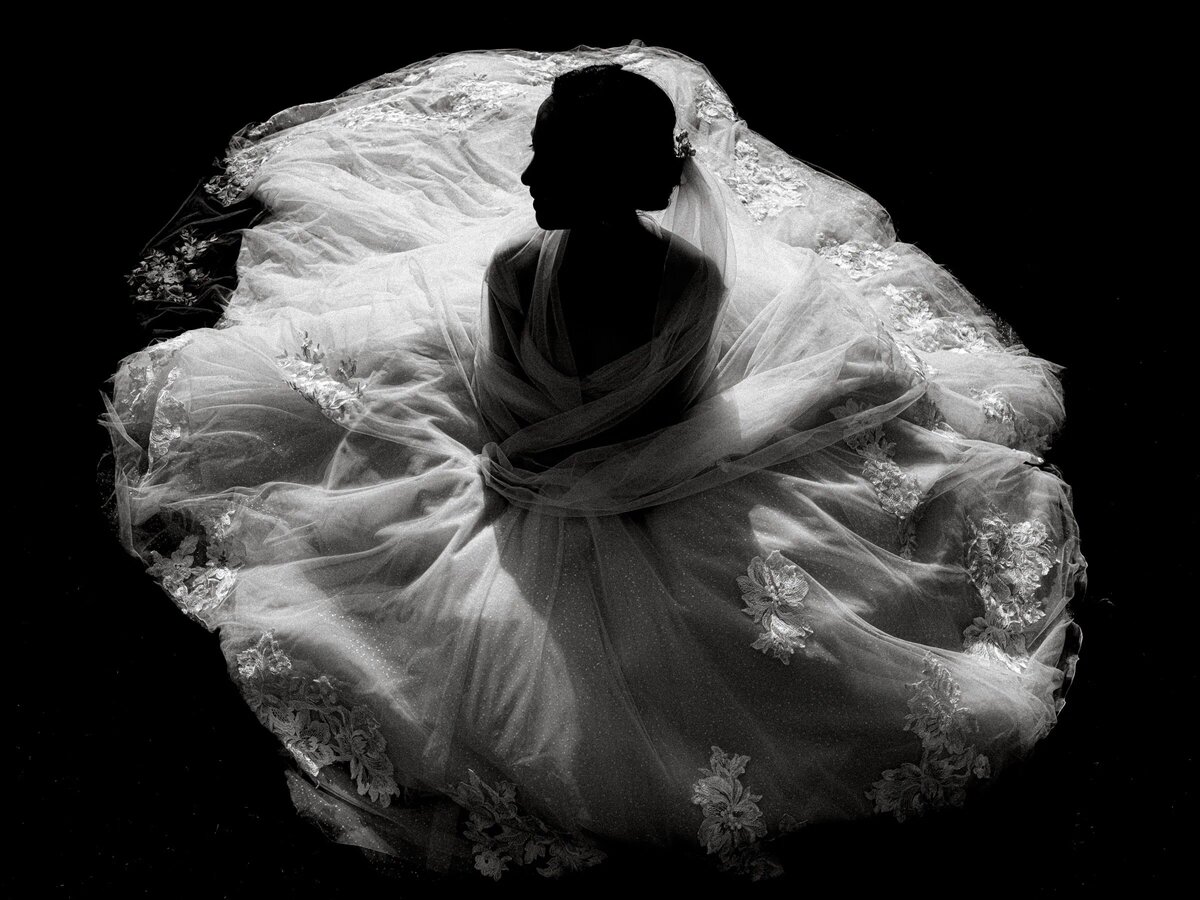 A bride seated on the floor in her voluminous wedding dress, her silhouette outlined by the ambient light