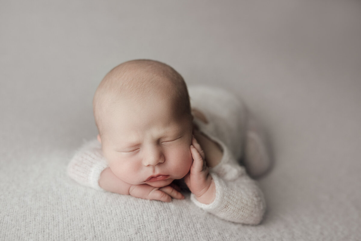 Studio newborn photography - sleeping baby on his belly with one hand under his chin, the other resting on his cheek. Baby is wearing a white knit onesie on a white blanket.