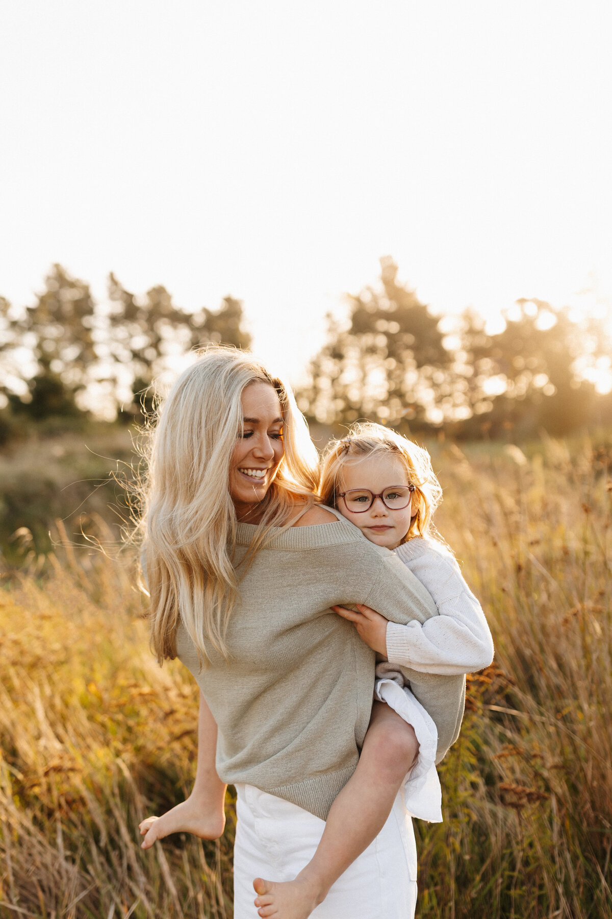 A mother gives her daughter a piggyback while being photographed at sunset in a field of long grass while lucy jane photographs them in a candid and documentary style in canterbury