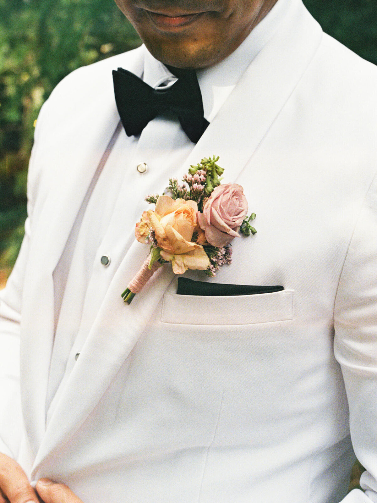 Groom in a white suit with a black bow tie and a pastel floral boutonniere on the lapel. The mood is elegant and formal against a blurred greenery backdrop.