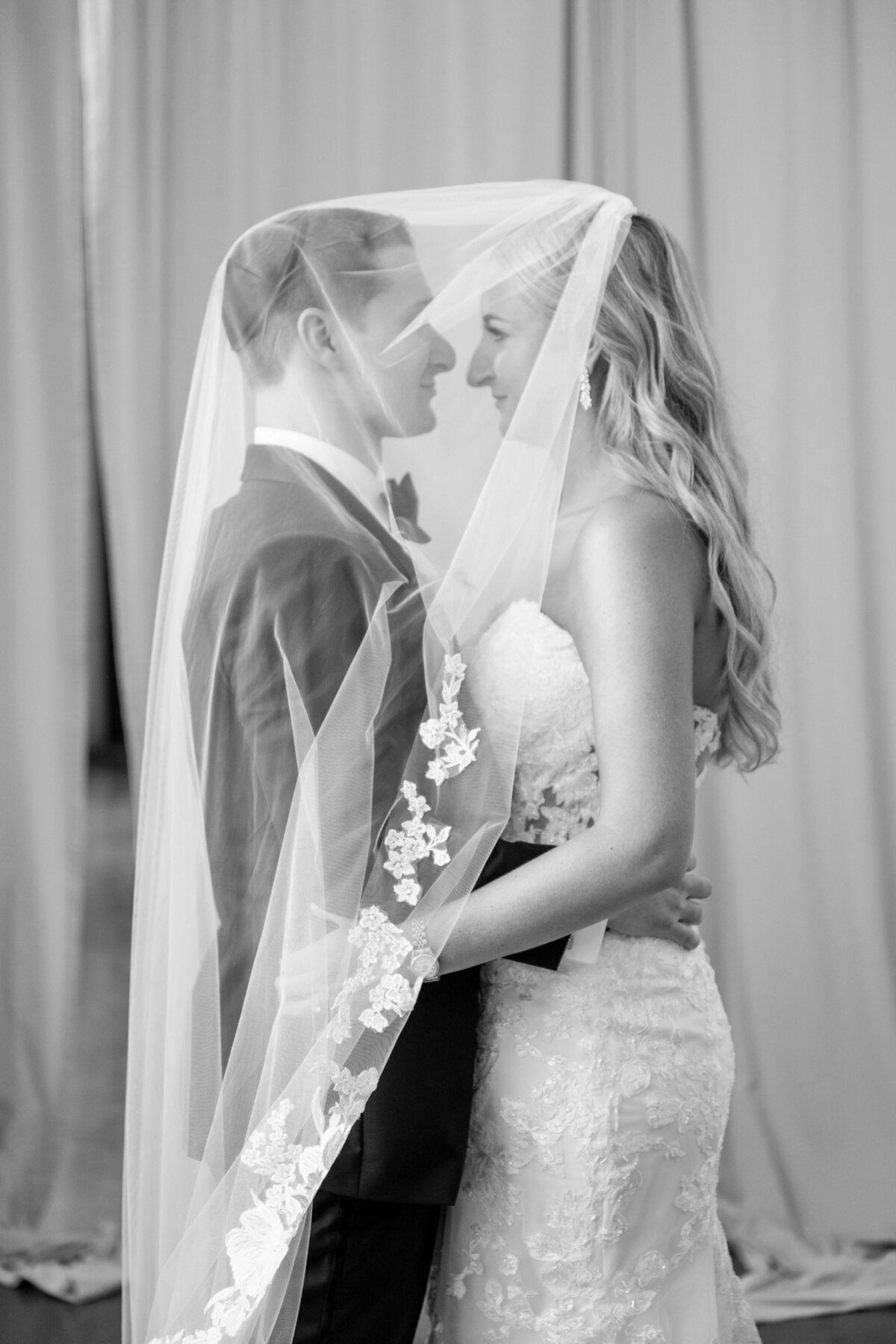 A black and white photo of a couple embracing under a bridal veil. They are facing each other and smiling. The bride is wearing a lace dress, and the groom is in a suit. The background consists of curtains.