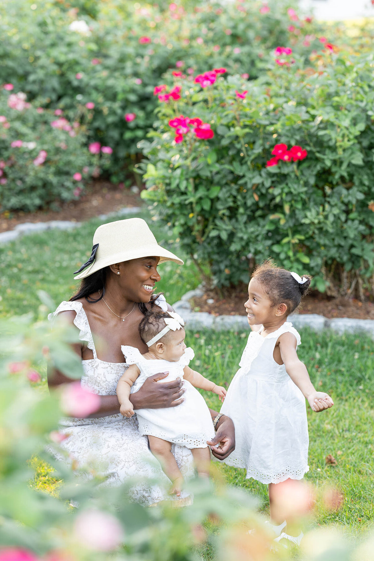 Woman wearing a straw hat and tan dress is kneeling down as she holds her baby girl on her lap while her toddler is standing in front of her with her arms stretched horizontally during their mommy and me session in a rose garden