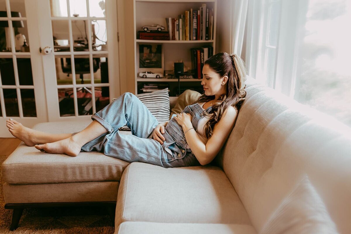 A woman in denim overalls relaxes on a beige sectional sofa, holding her stomach. She is sitting in a well-lit room with bookshelves and closed glass-paneled doors in the background.