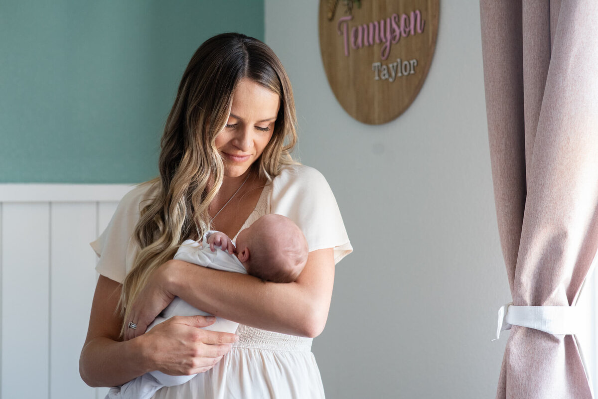 A smiling mother stands in a nursery window smiling down at her newborn in her arms