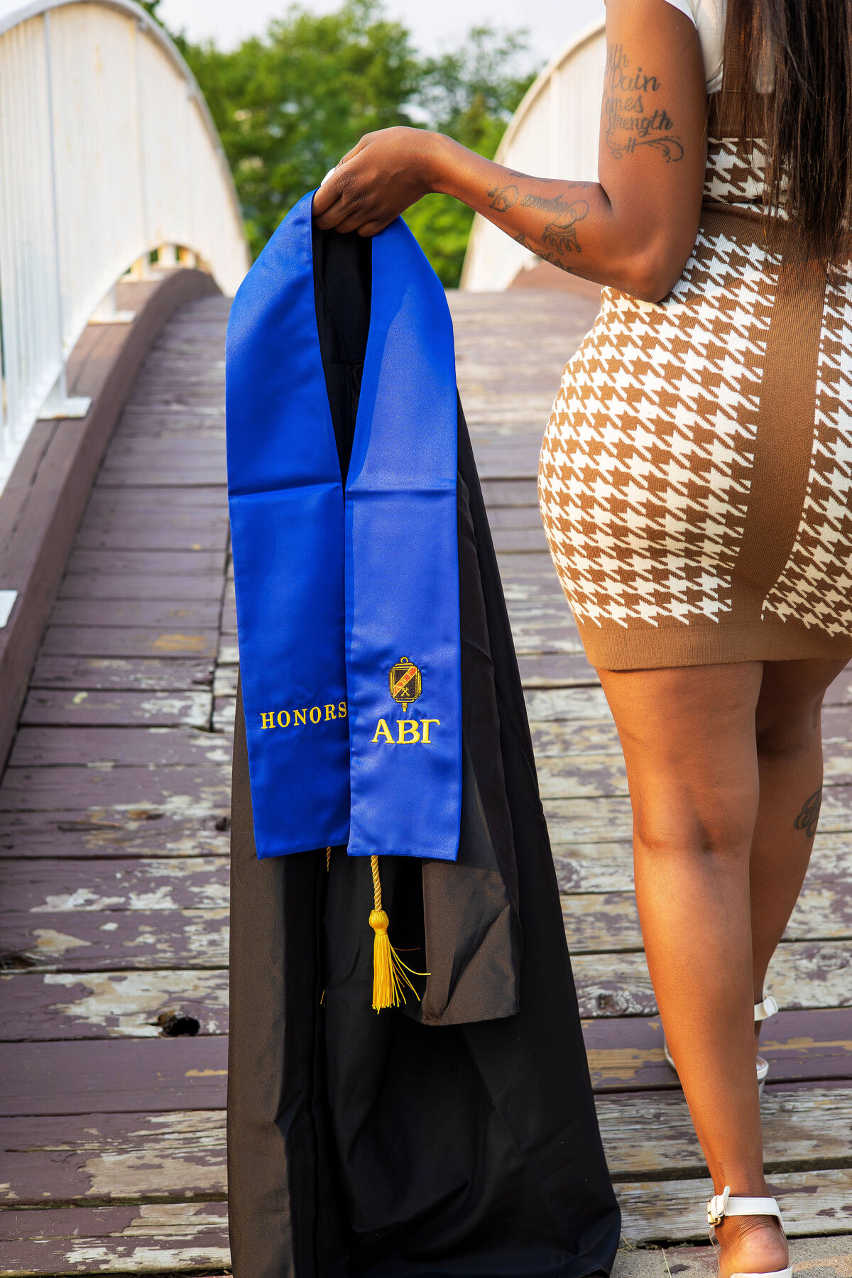 Woman holding graduation gown, shash, and honors with back turned in front of a bridge