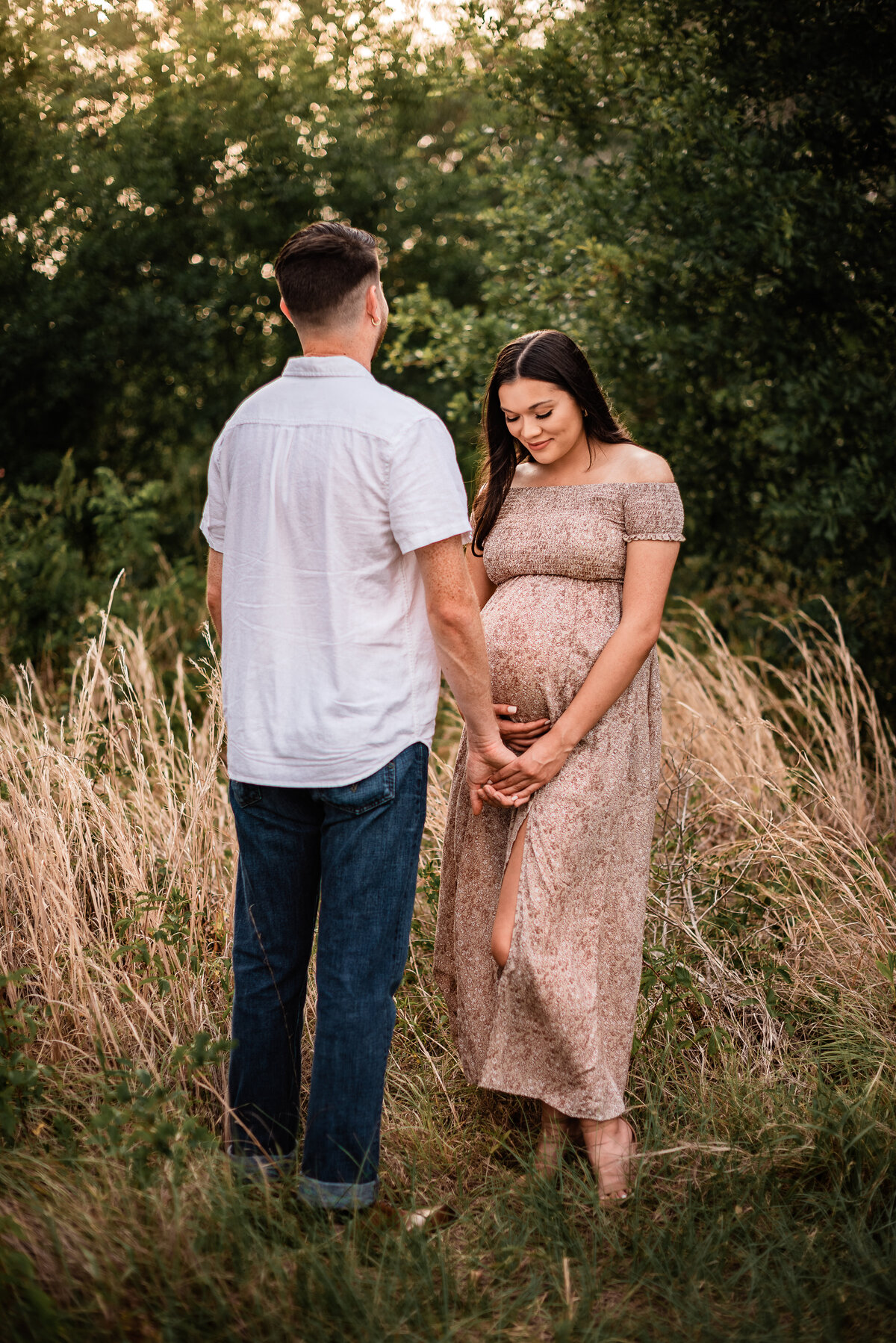 A Houston area mom to be holds her boyfriend's hand and looks at her belly while standing in a field of golden grass.