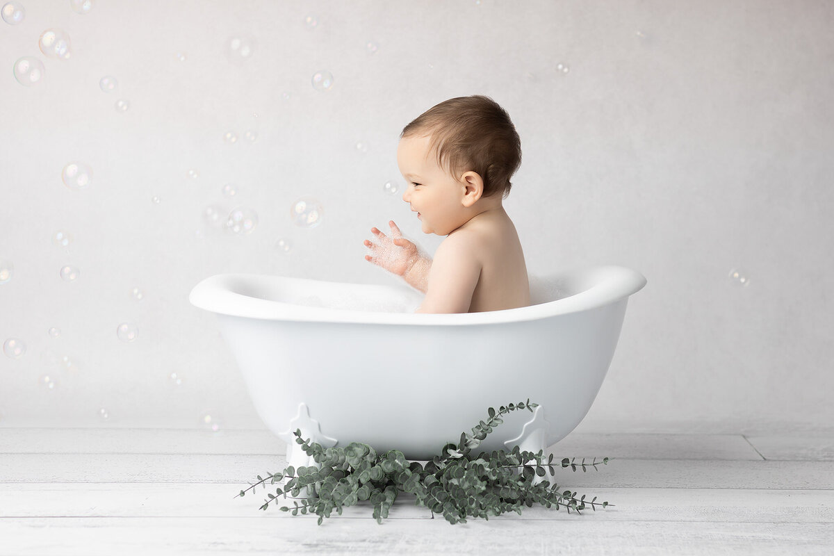 Little boy splashes with delight in a bubble-filled bath set inside a white clawfoot tub accented with eucalyptus, captured during a DFW milestone photography session.