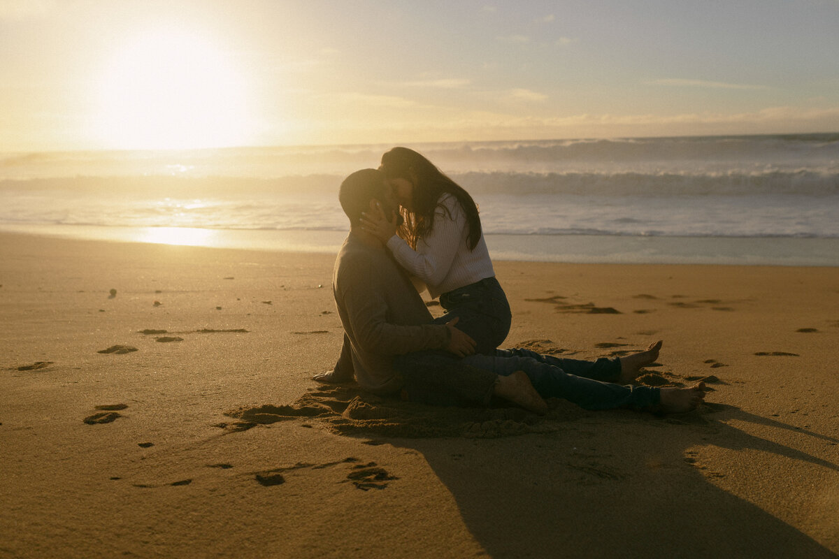 Girl sitting on top of guy on sand on beach on California coast during sunset in front of ocean about to kiss