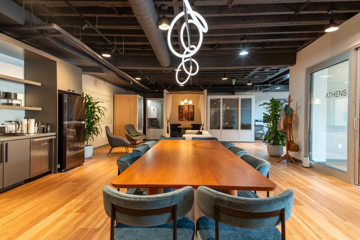 Large communal space with a long wood table, blue upholstered chairs, a kitchenette, lounge areas, and sculptural modern lighting overhead.