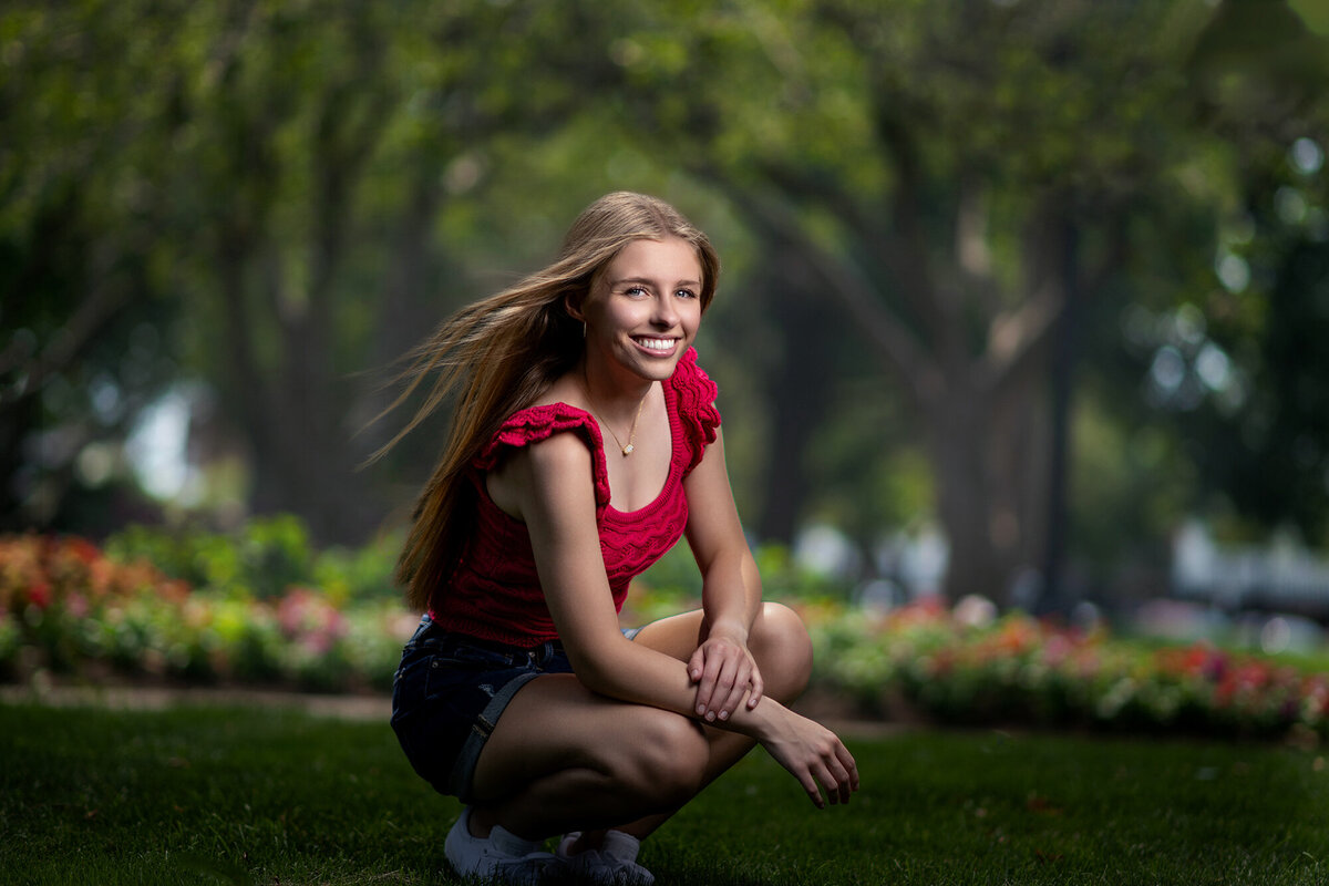 A woman kneels in a garden in a red top and jean shorts with her blonde hair blowing in the wind
