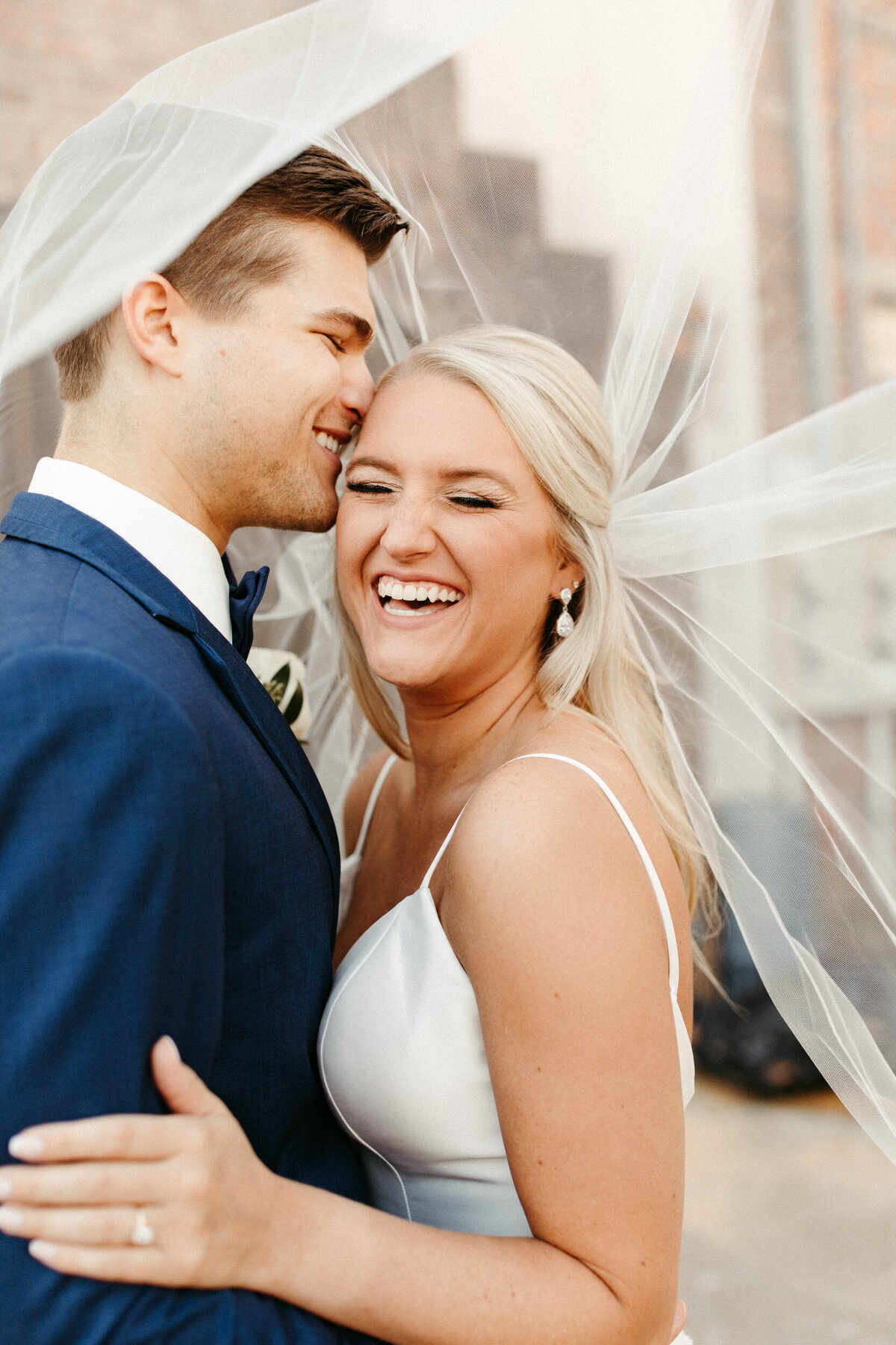 Groom in blue suit laughing and hugging his giggly bride as the wind blows her veil over them