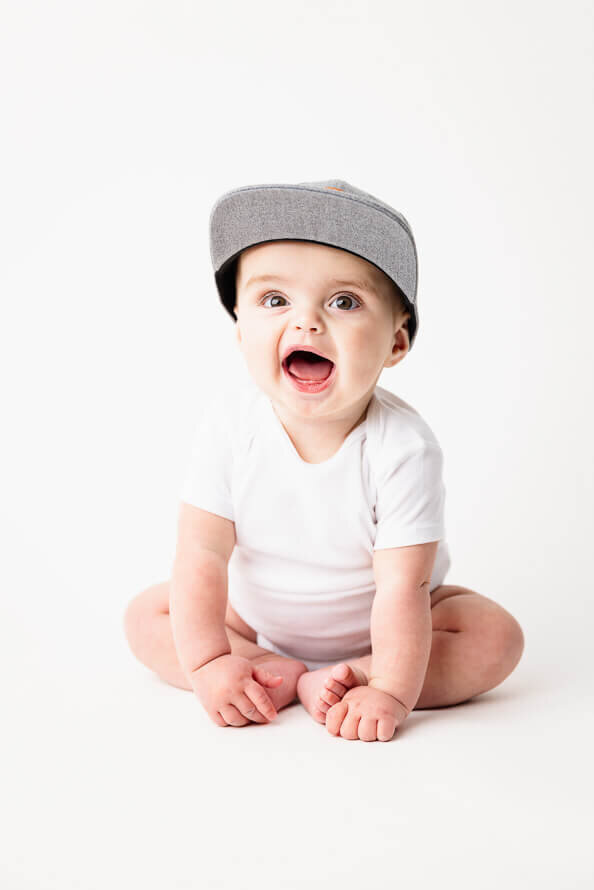 One year old boy in hat smiles at photographer in a white studio.