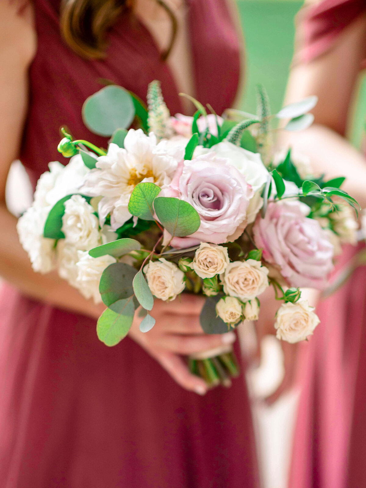 A woman in a maroon dress holding a bouquet of flowers, including pink roses, white dahlias, and eucalyptus leaves. The background is blurred, focusing on the floral arrangement.