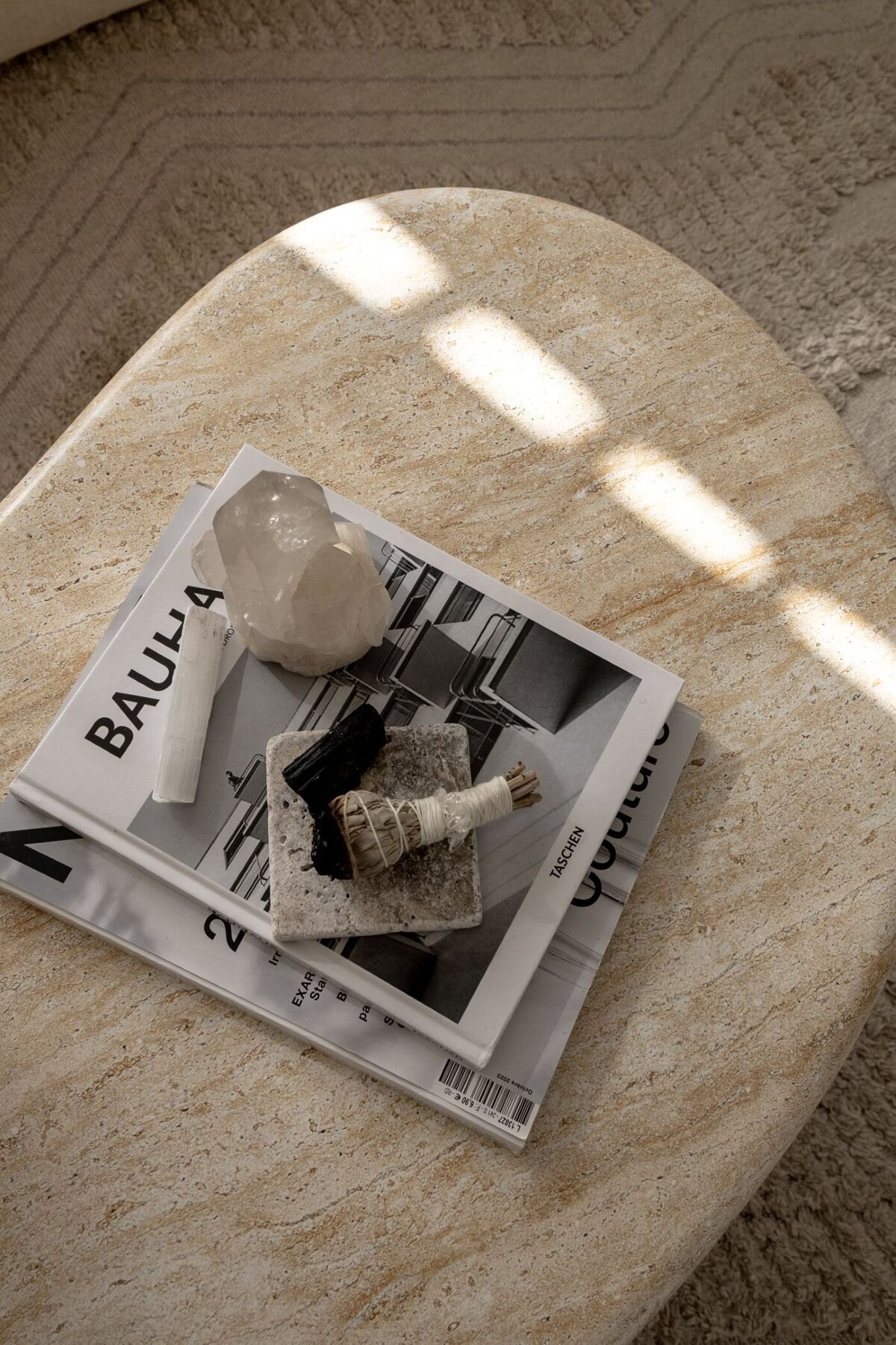 A travertine coffee table viewed from above, adorned with a Bauhaus design book, a magazine, a crystal, and sage. A white wool patterned rug provides a soft contrast in the background.