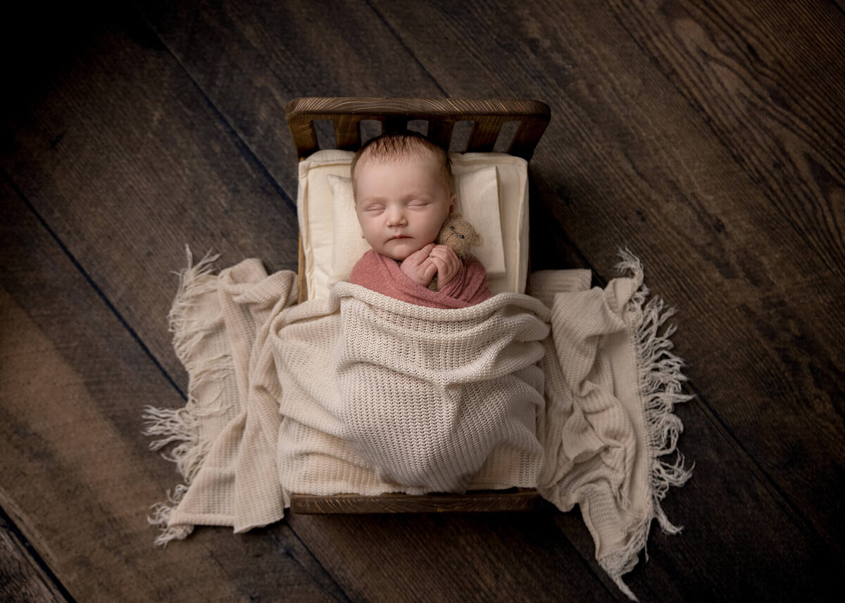 newborn baby asleep in a tiny wooden bed with a cream blanket and holding a tiny teddy bear