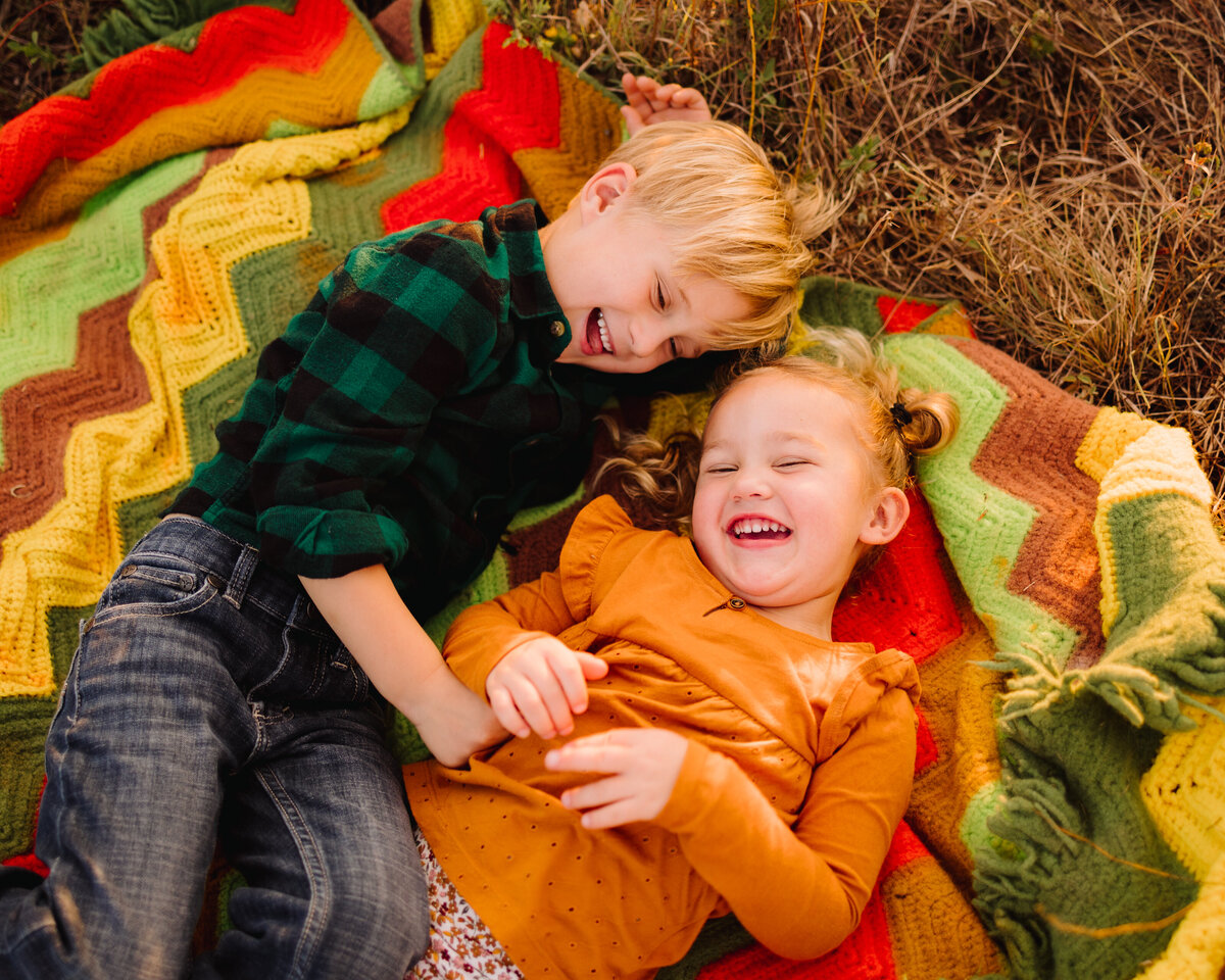 Photo of brothers smiling on a blanket of various colors green, orange, red and brown. The blonde girl has her eyes closed and is wearing an orange shirt, and the boy is wearing a green plaid shirt and jeans.Photo of brothers smiling on a blanket of various colors green, orange, red and brown. The blonde girl has her eyes closed and is wearing an orange shirt, and the boy is wearing a green plaid shirt and jeans.