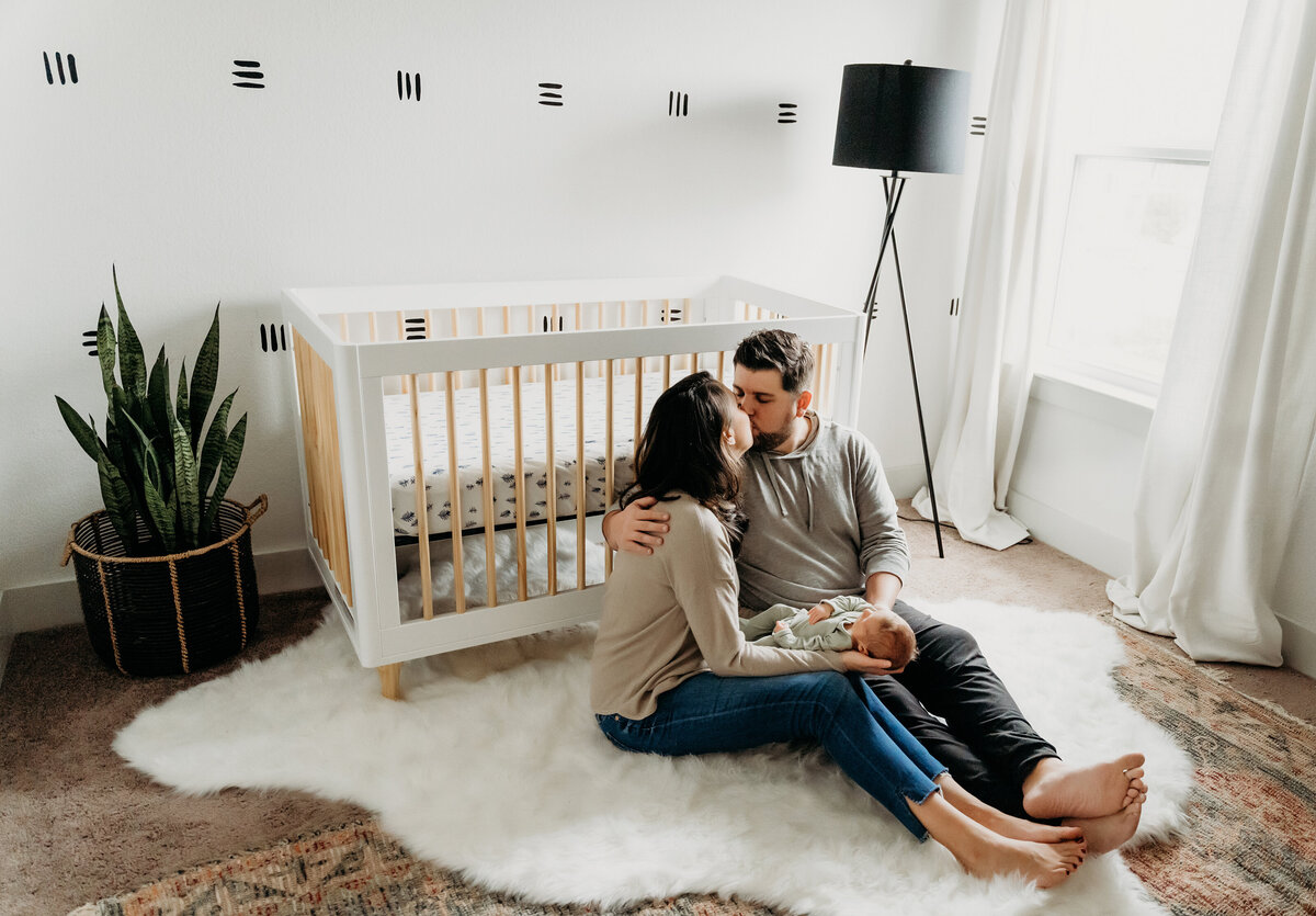 Newborn Photographer,  a young mother and father sit in the nursery floor holding their new baby, they kiss