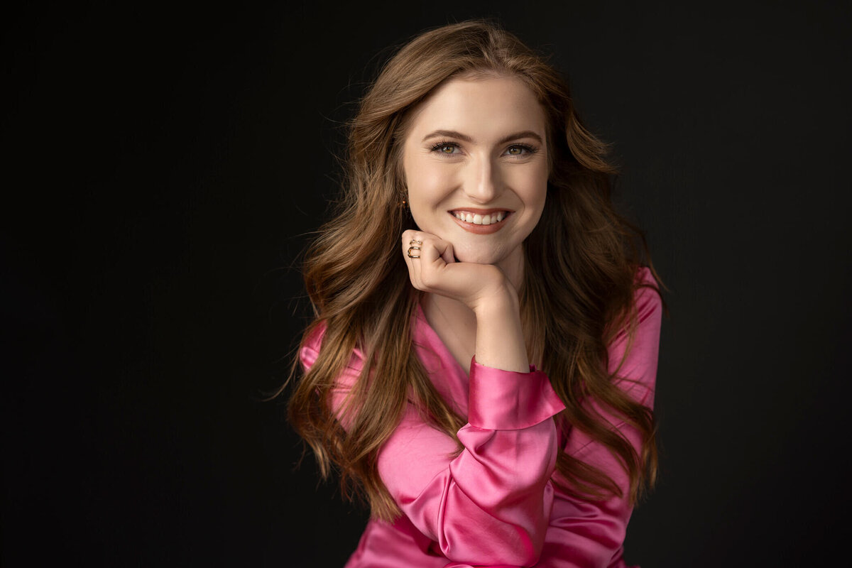 Young woman in a pink satin blouse, smiling with her chin resting on her hand.
