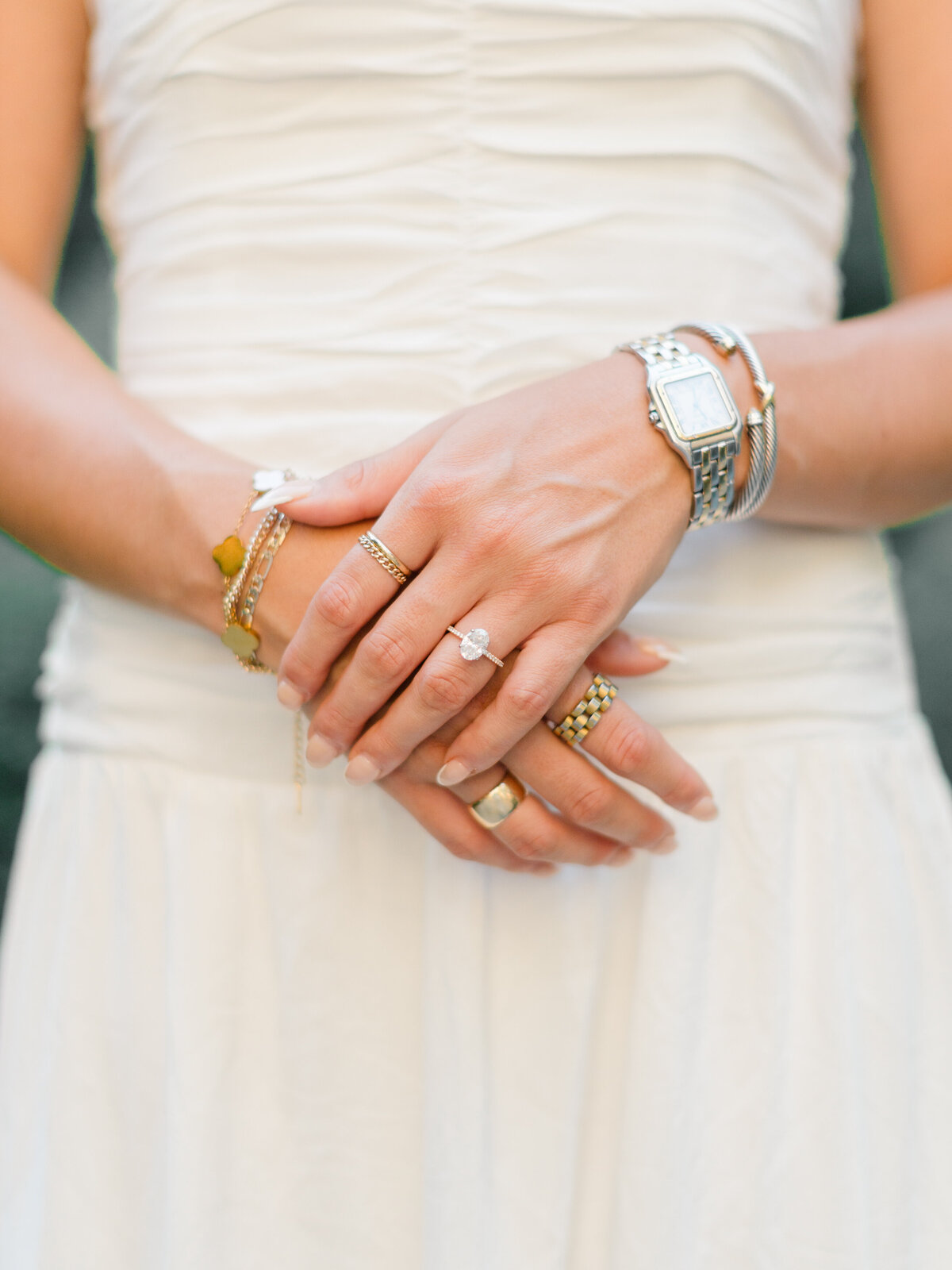 A close-up of a bride-to-be’s hands gently clasped in front of her white dress, showcasing her engagement ring and a curated mix of gold and silver jewelry. This elegant detail shot, taken in Rittenhouse Square, was captured by Elizabeth Kane Photography, a Luxury Philadelphia Wedding Photographer.