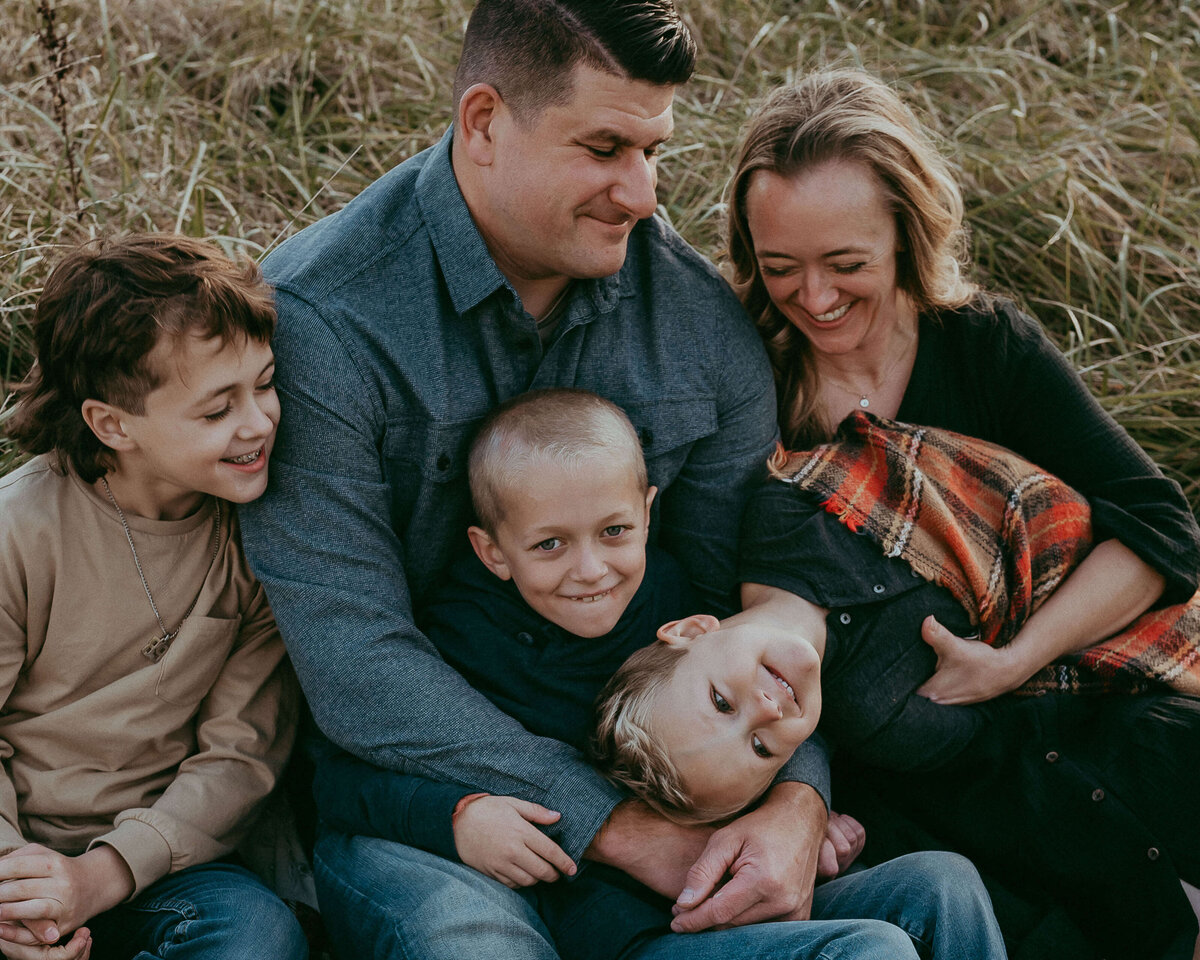 A mom and dad sit on a blanket during fall with their three wild boys, laughing and giggling while wearing a stylish neutral color palette that coordinates perfectly with the bottom tones of the tall grasses they're sitting in.
