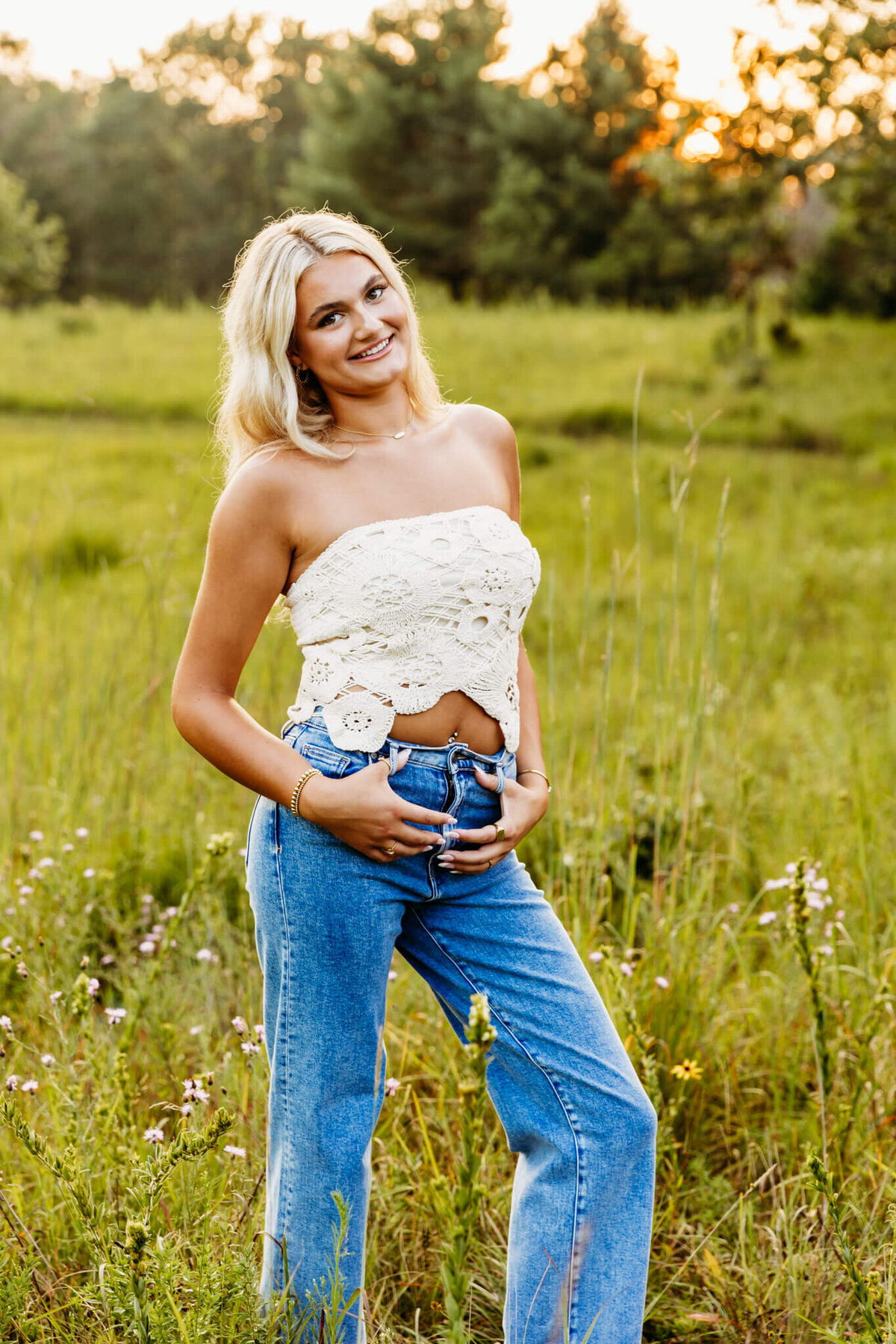 pretty high school girl in a white lace top and jeans standing in a beautiful field and smiling captured by Ashley Kalbus.