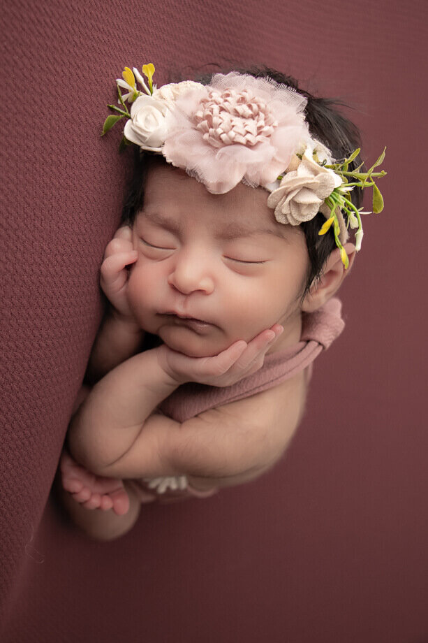 A baby is in the timber pose on a rich burgundy backdrop.  She is wearing a mauve romper with a matching floral headband
