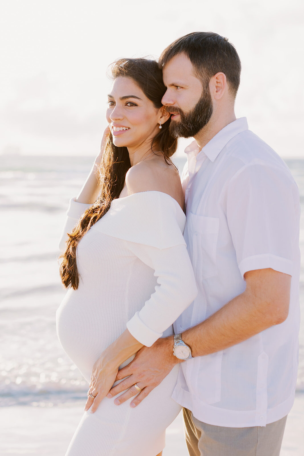 A glowing mother-to-be leans into her partner as he lovingly embraces her baby bump, bathed in warm sunlight on a Florida beach. Captured by Claudia Amalia Maternity Photography.