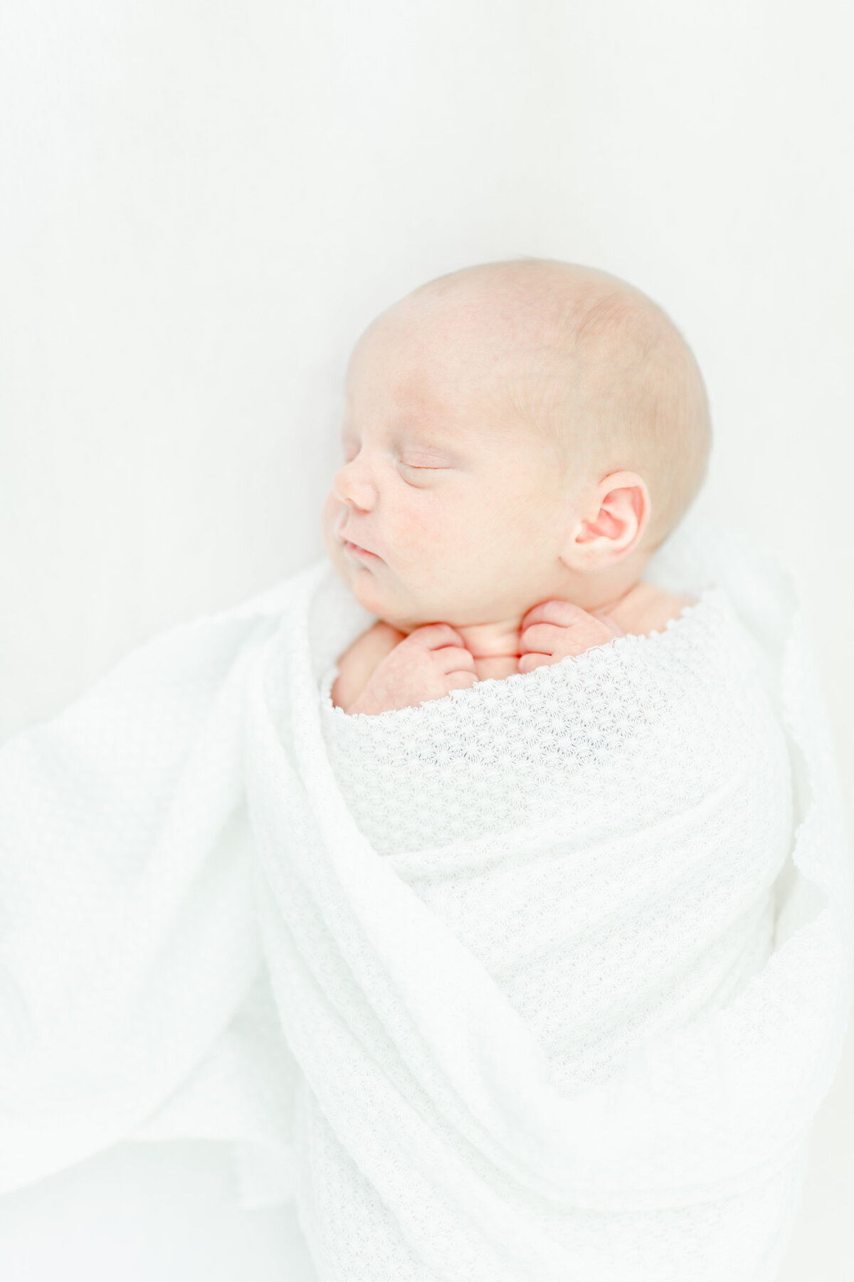 Newborn baby wrapped in a white blanket with his tiny hand under his chin. Newborn photo taken in a studio in New Jersey