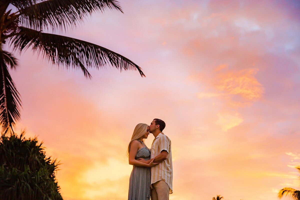 Man kisses his wife on the forehead as she wears an aqua Kasia Kulenty gown at sunrise for their engagement session in Maui