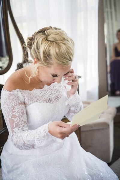 A bride sits in her wedding dress, wiping a tear from her eye as she reads a letter from her groom.