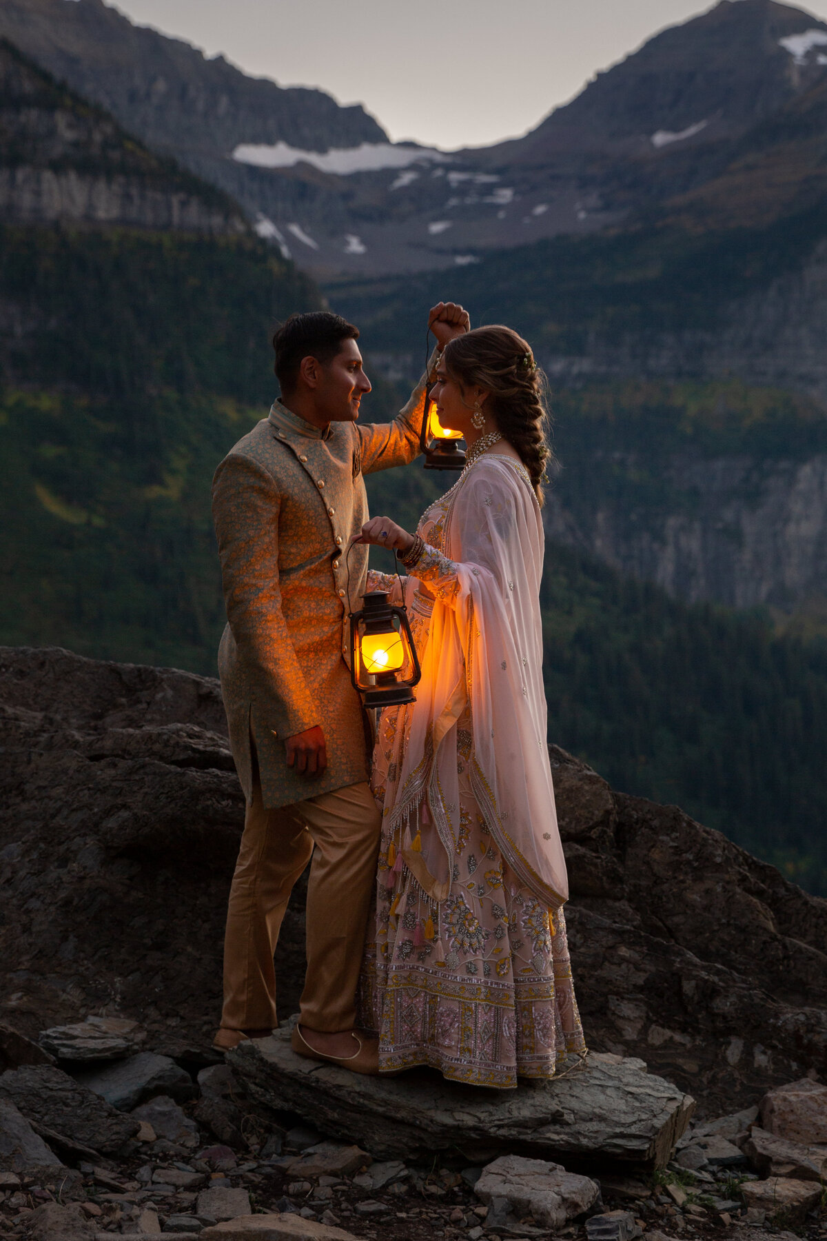 A bride and groom wearing Indian wedding attire stand facing each other on a rock in Glacier National Park, holding lanterns up to provide light.