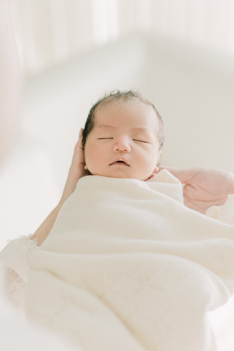 ママに抱かれ、白いブランケットにくるまれて眠っている可愛らしい赤ちゃんのナチュラル・ニューボーンフォト。