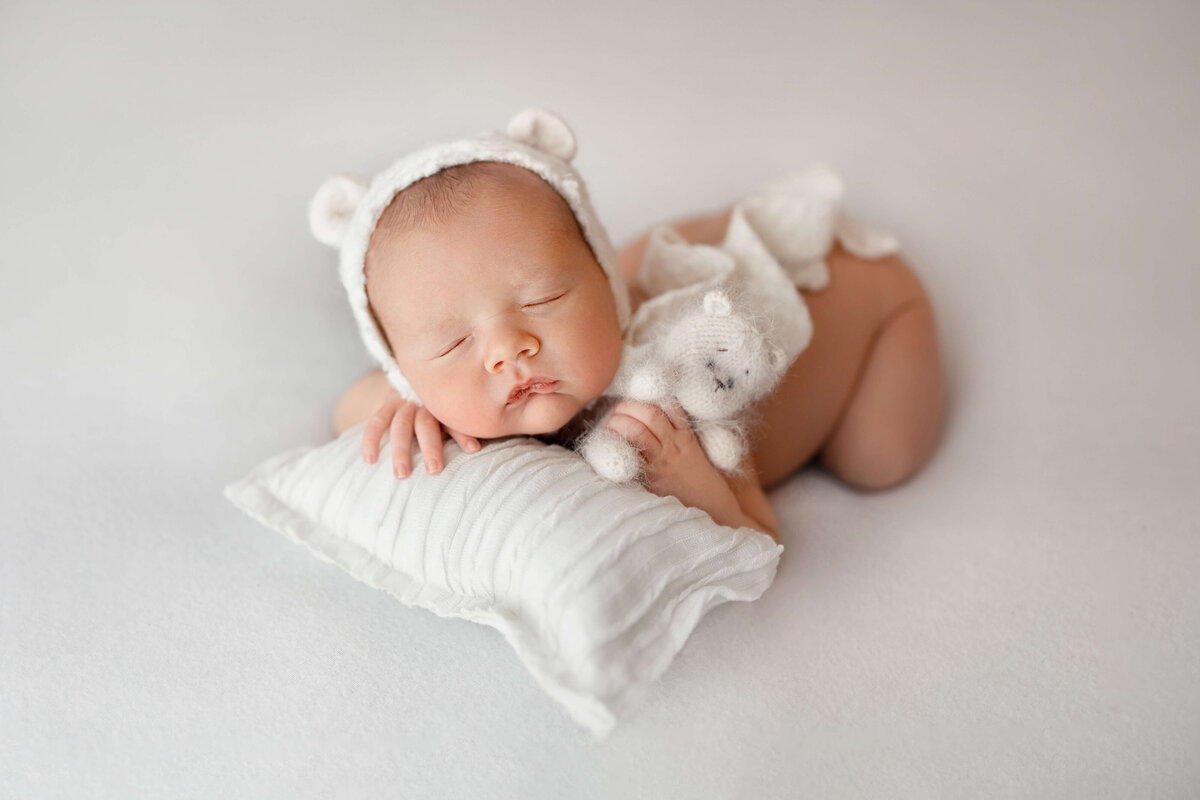 baby boy with a white bear hat on with his head and hand on a white pillow  holding a white bear at his  newborn photography session with a photographer at  a mclean va photo studio