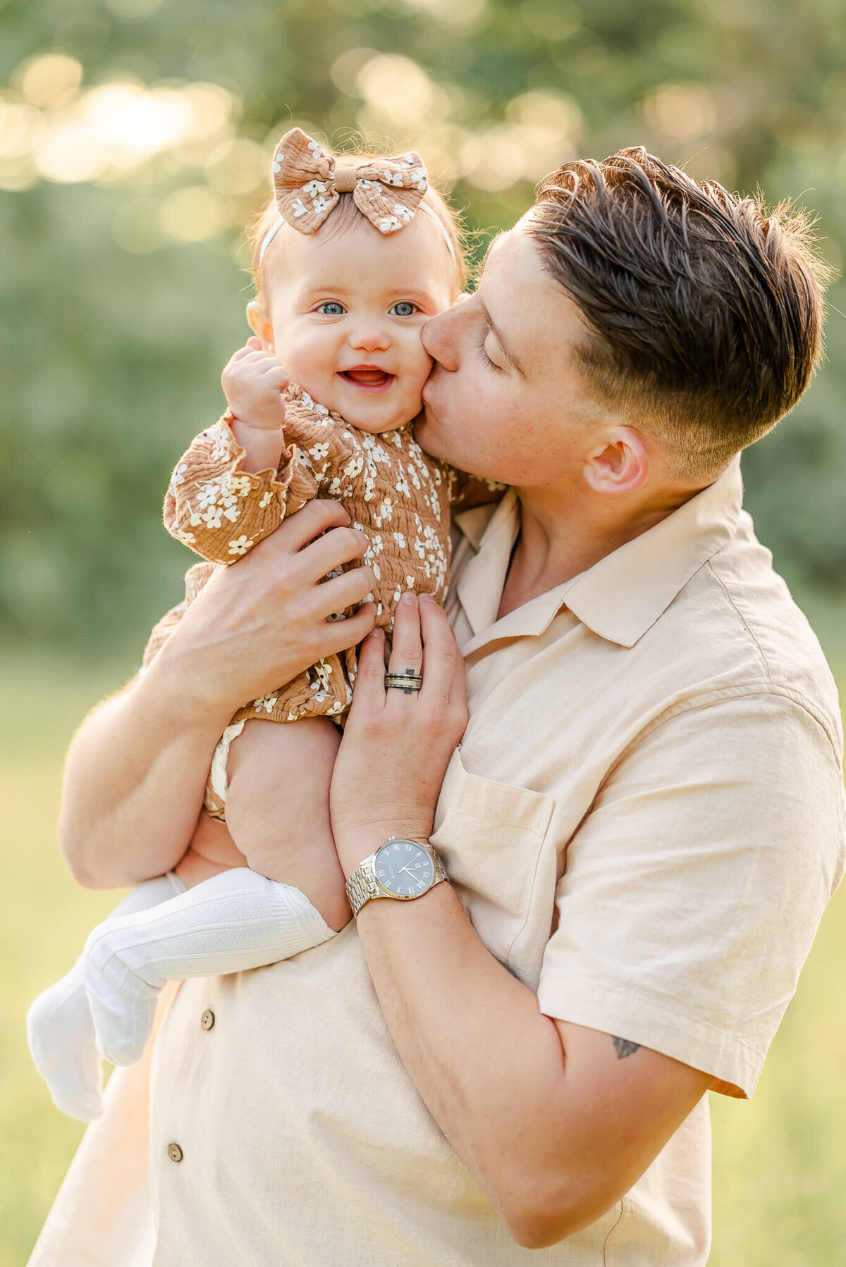 A man in an off-white button down, kisses his baby daughter on the cheek. The baby is wearing a brown floral romper and matching bow.