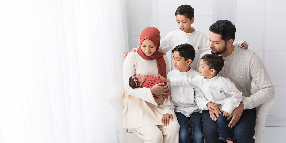 Parents and three young boys gathered around their newborn baby girl as the mother holds her, all dressed in soft neutral tones during a family portrait.