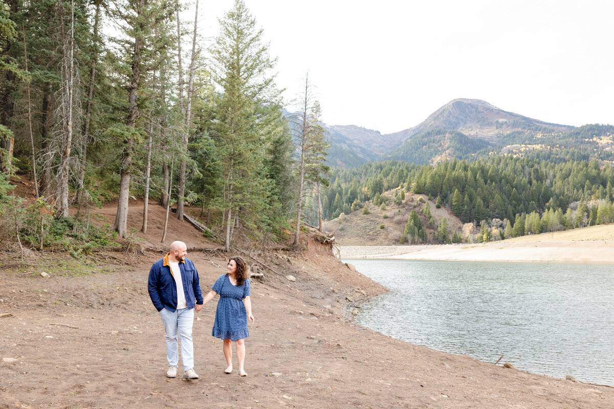 A couple walks hand in hand along the shoreline of Tibble Fork Reservoir, surrounded by towering pine trees and majestic mountains. The woman in a blue dress looks up at her partner in admiration as they stroll through nature.