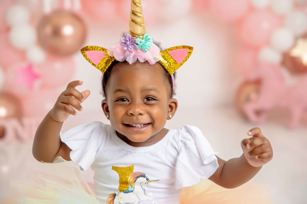 A joyful toddler, captured by a Jacksonville cake smash photographer, wears a unicorn-themed headband with golden ears and horn. Dressed in a white shirt featuring a unicorn print and colorful tutu, the scene is enchantingly set with pink and white balloons, perfect for cake smash photography.