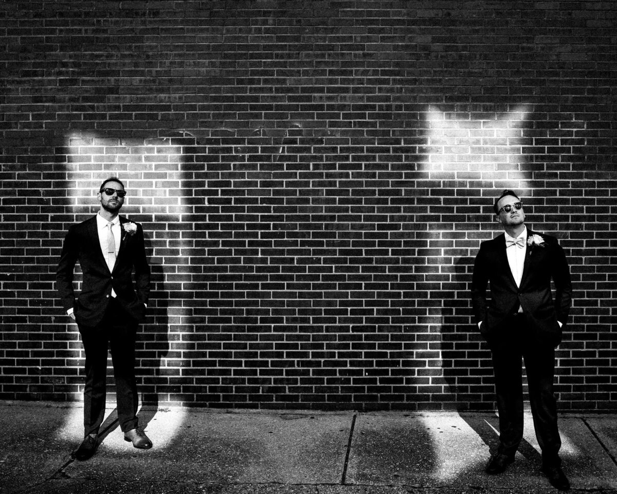 Two men in formal attire standing apart against a brick wall with wing-like light reflections over their heads