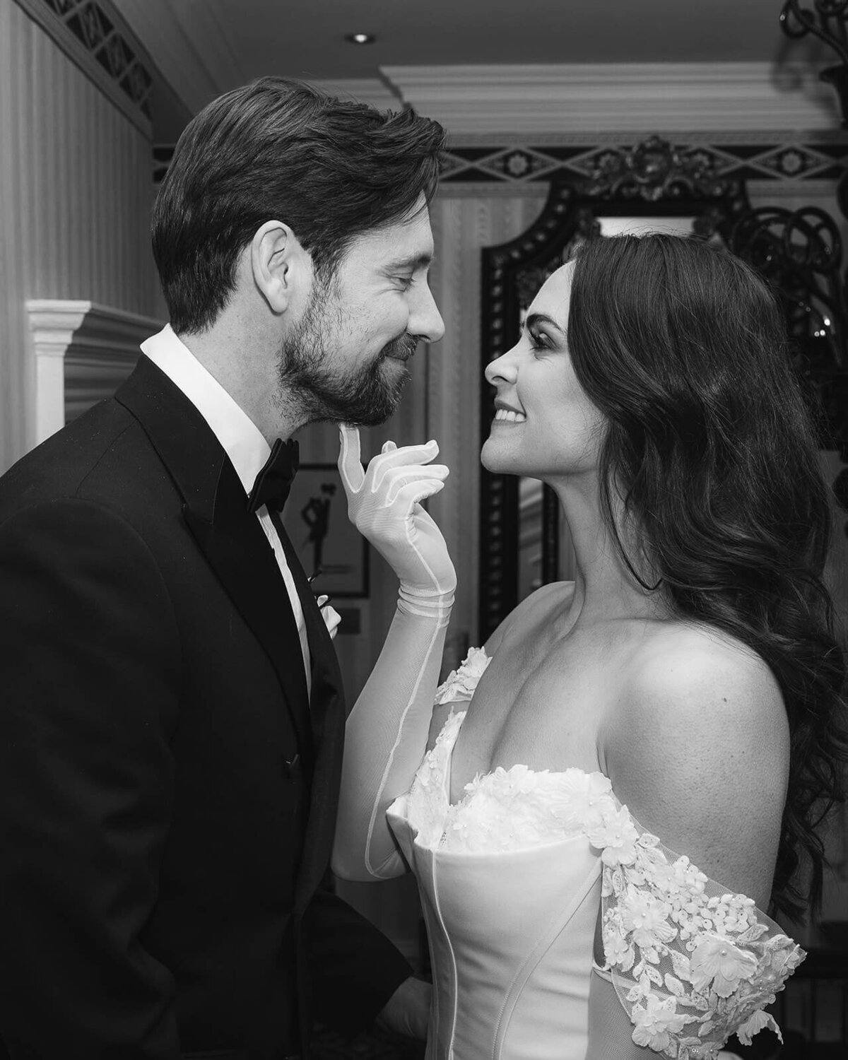 A bride wearing gloves tickles her husbands chin as they stare eye to eye